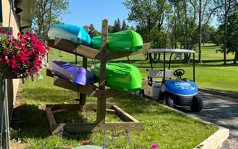 Picture of a kayak rack with four kayaks next to a hanging flower basket, golf cart at a golf course
